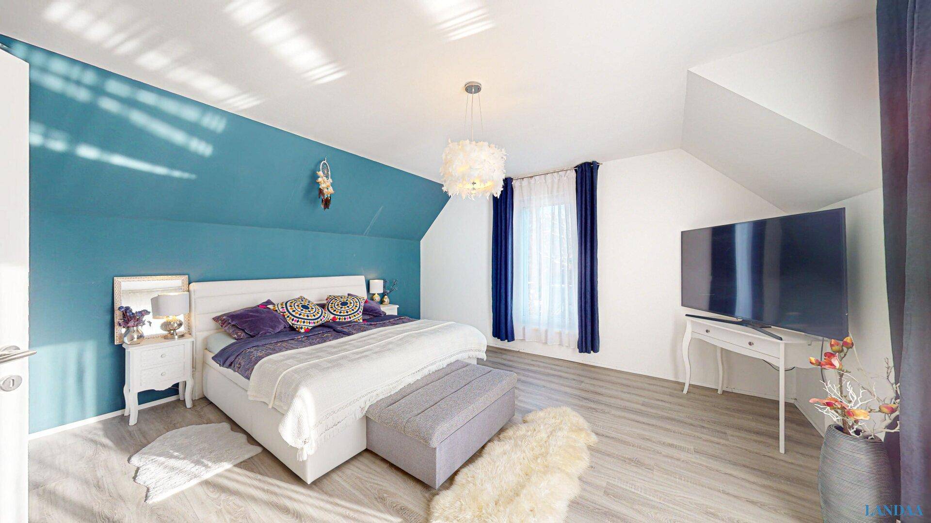 Helles Schlafzimmer mit Dachschräge, Fernseher und blauer Wandgestaltung.