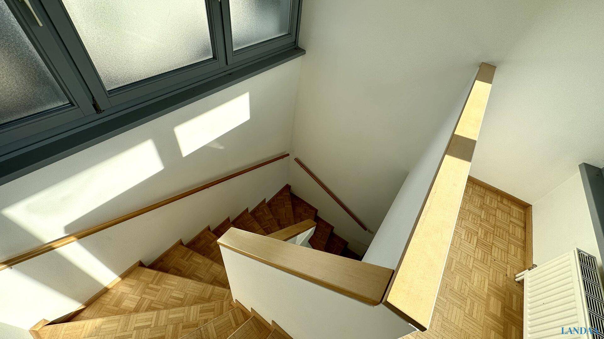 Helles Treppenhaus mit Holzstufen und Parkettboden, gut beleuchtet durch Fenster.