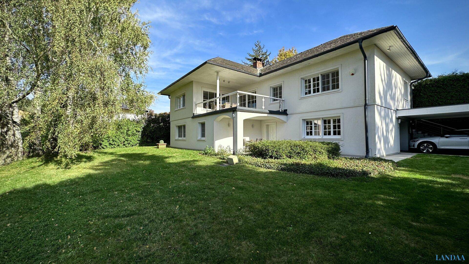 Ansicht des Hauses mit weitläufigem Garten, Balkon und Carport unter blauem Himmel.
