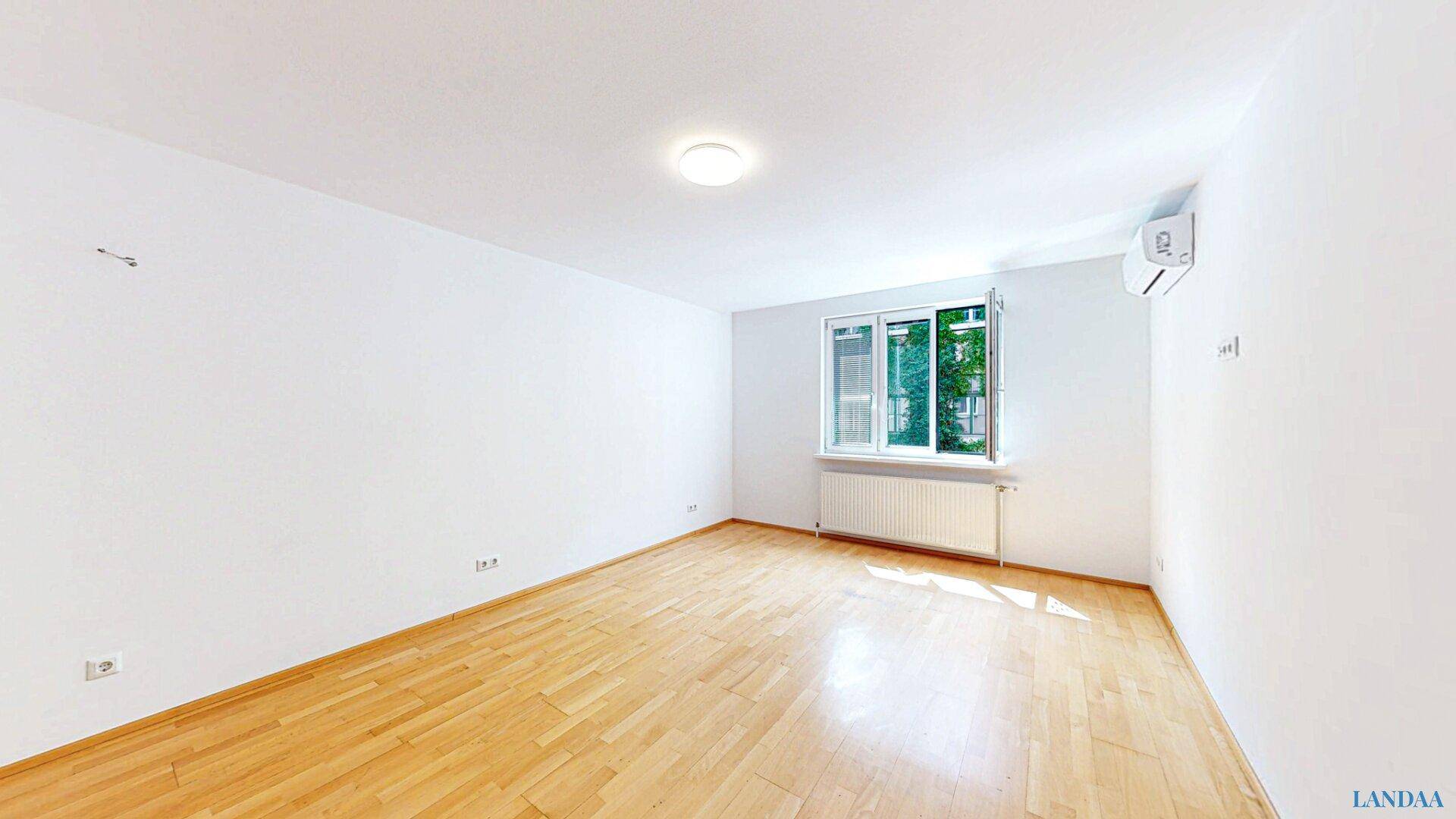 Leeres Zimmer mit hellem Holzboden, großen Fenstern und Klimaanlage.