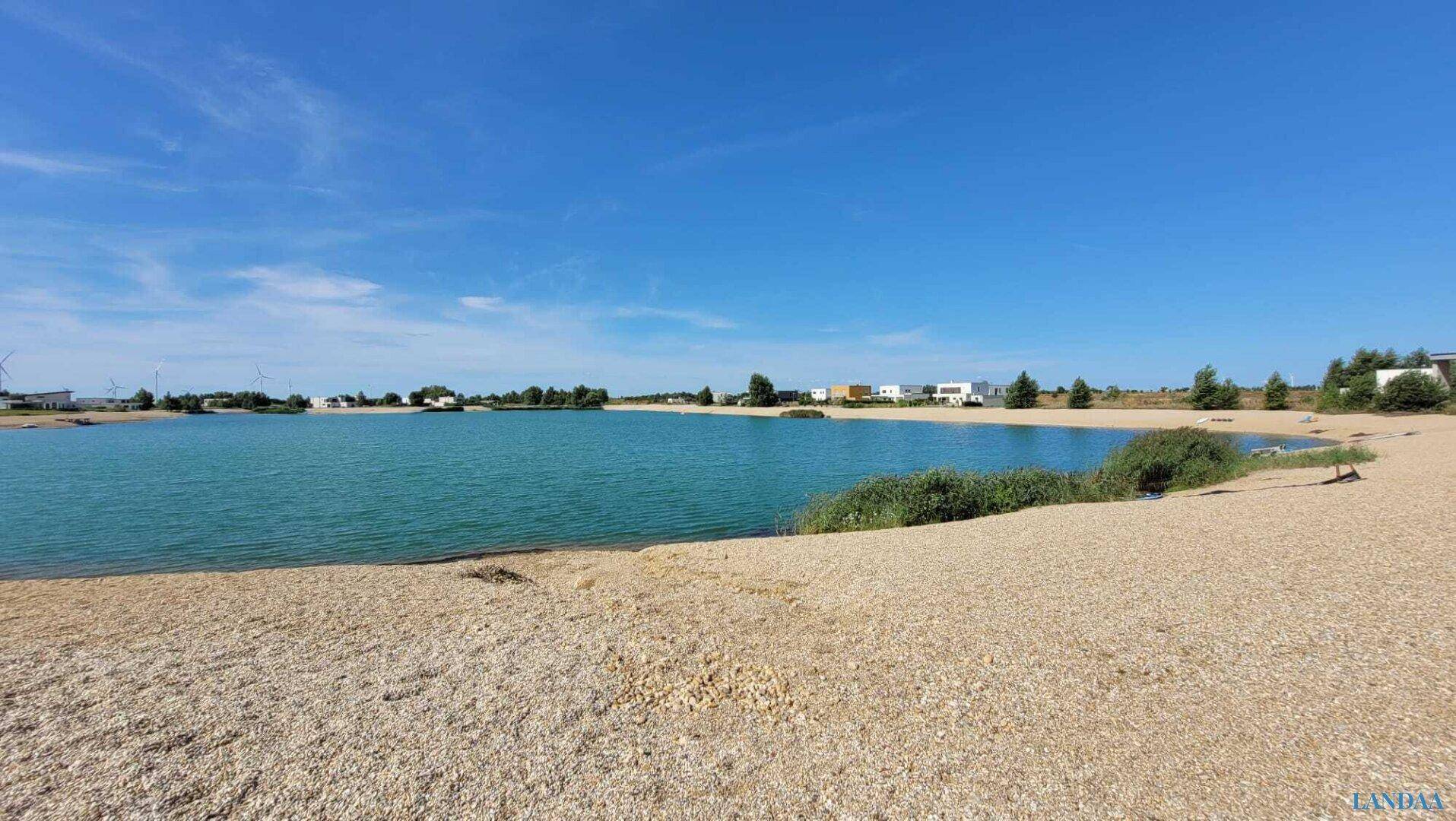 Weitläufiger Blick auf den See mit Sandstrand und umliegender Natur.
