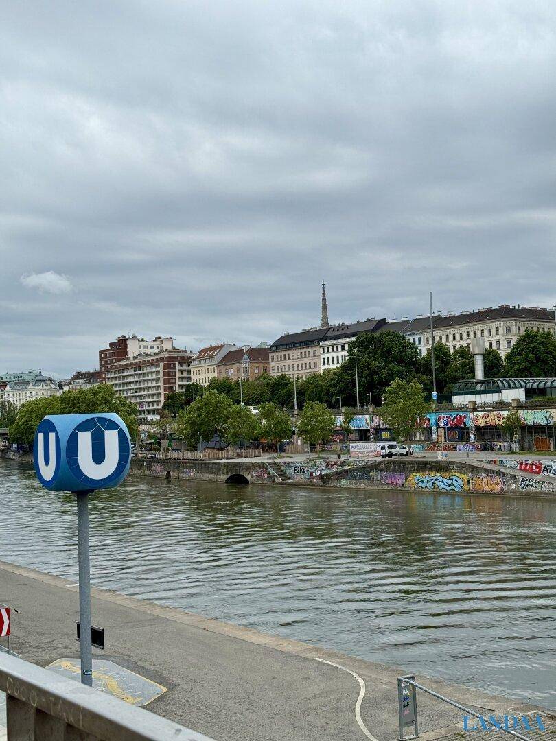 Panoramablick auf einen Fluss mit Stadtkulisse und U-Bahn-Station unter bewölktem Himmel.