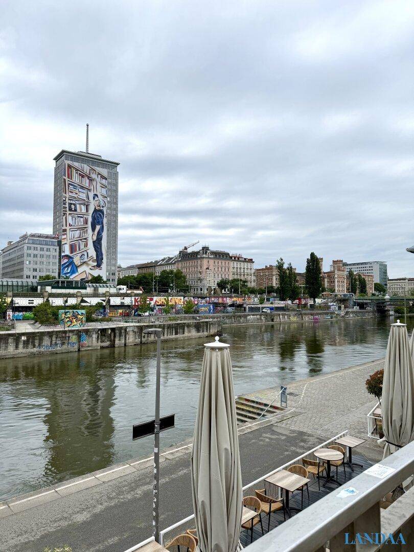 Blick auf einen Fluss mit Stadtkulisse und einem Café am Ufer.