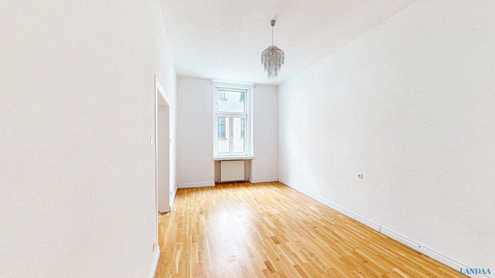 Leeres Zimmer mit Holzboden, weiß gestrichenen Wänden und einem Kronleuchter.