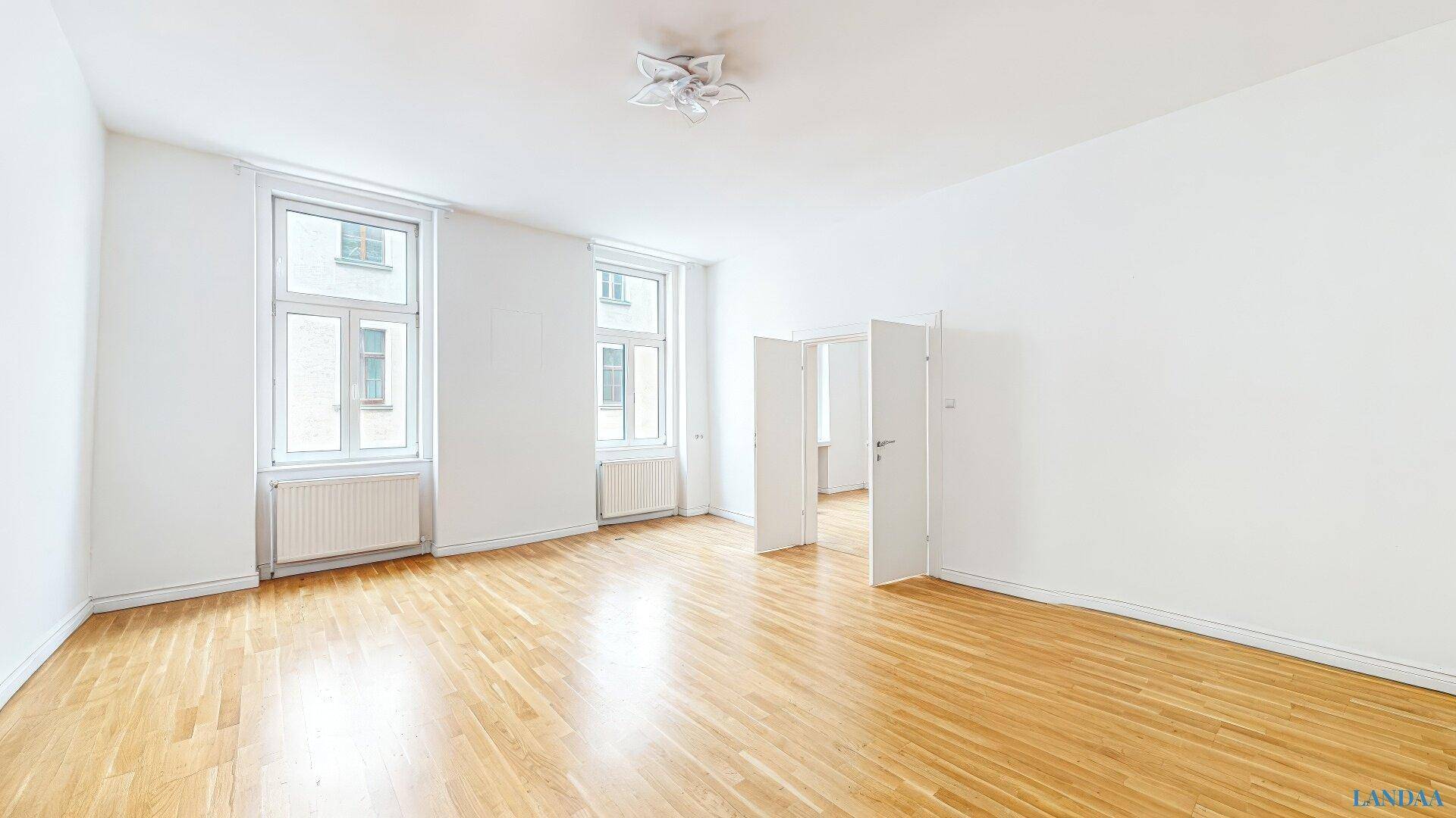 Geräumiges, unmöbliertes Wohnzimmer mit Holzboden und großen Fenstern.