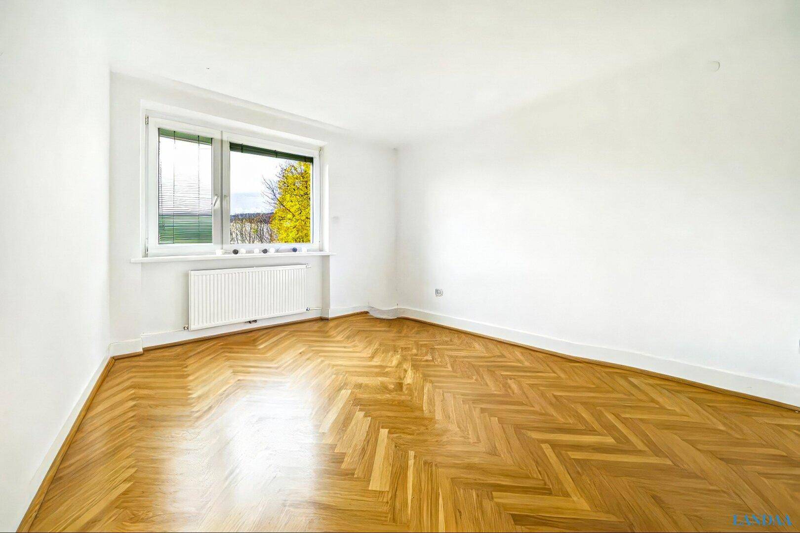 Heller, leerer Raum mit Fischgrätparkett, Heizung und grünem Ausblick.