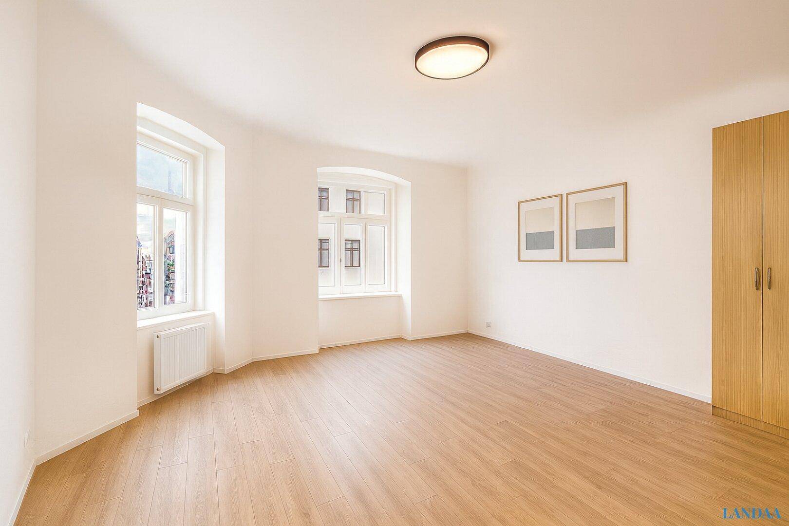 Leeres Zimmer mit Laminatboden, weißen Wänden und zwei Fenstern, bereit zur individuellen Gestaltung.