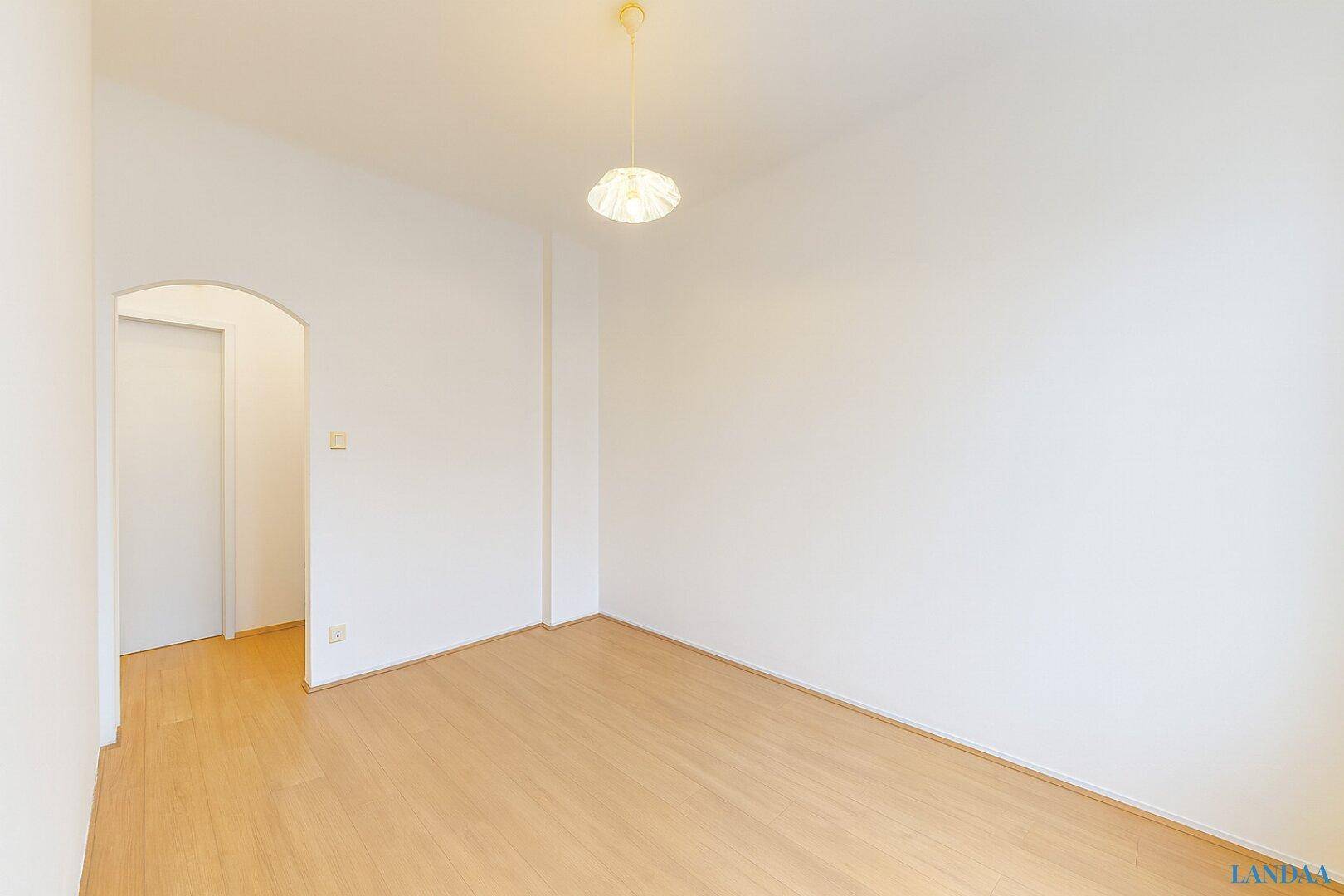 Leerer Raum mit hellem Holzboden und weiß gestrichenen Wänden, mit einem Bogendurchgang.