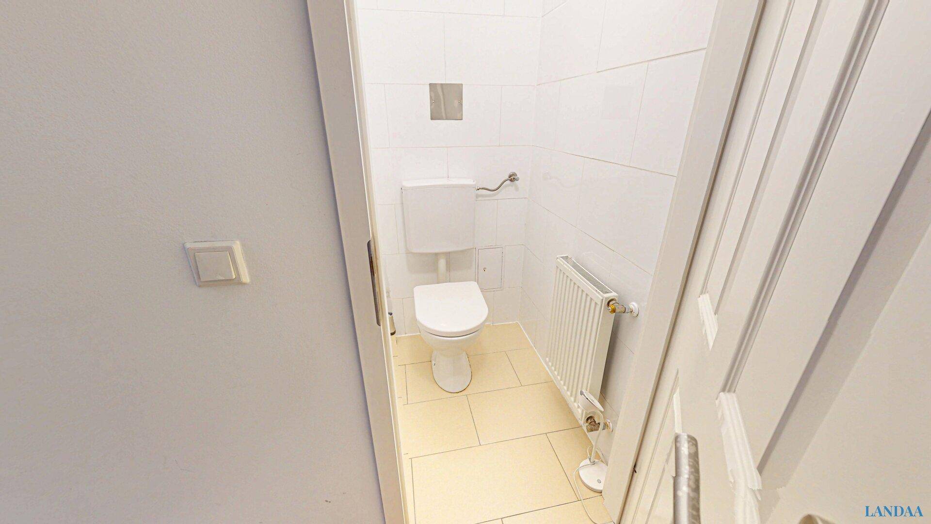 Separates WC mit Toilette, Heizkörper und hellen Fliesen an Wänden und Boden.