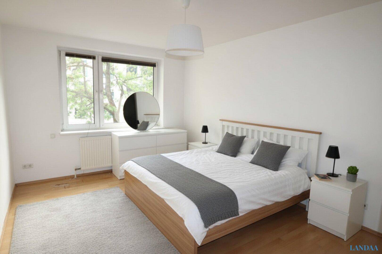 Helles Schlafzimmer mit Doppelbett, Kommode und großem Fenster.