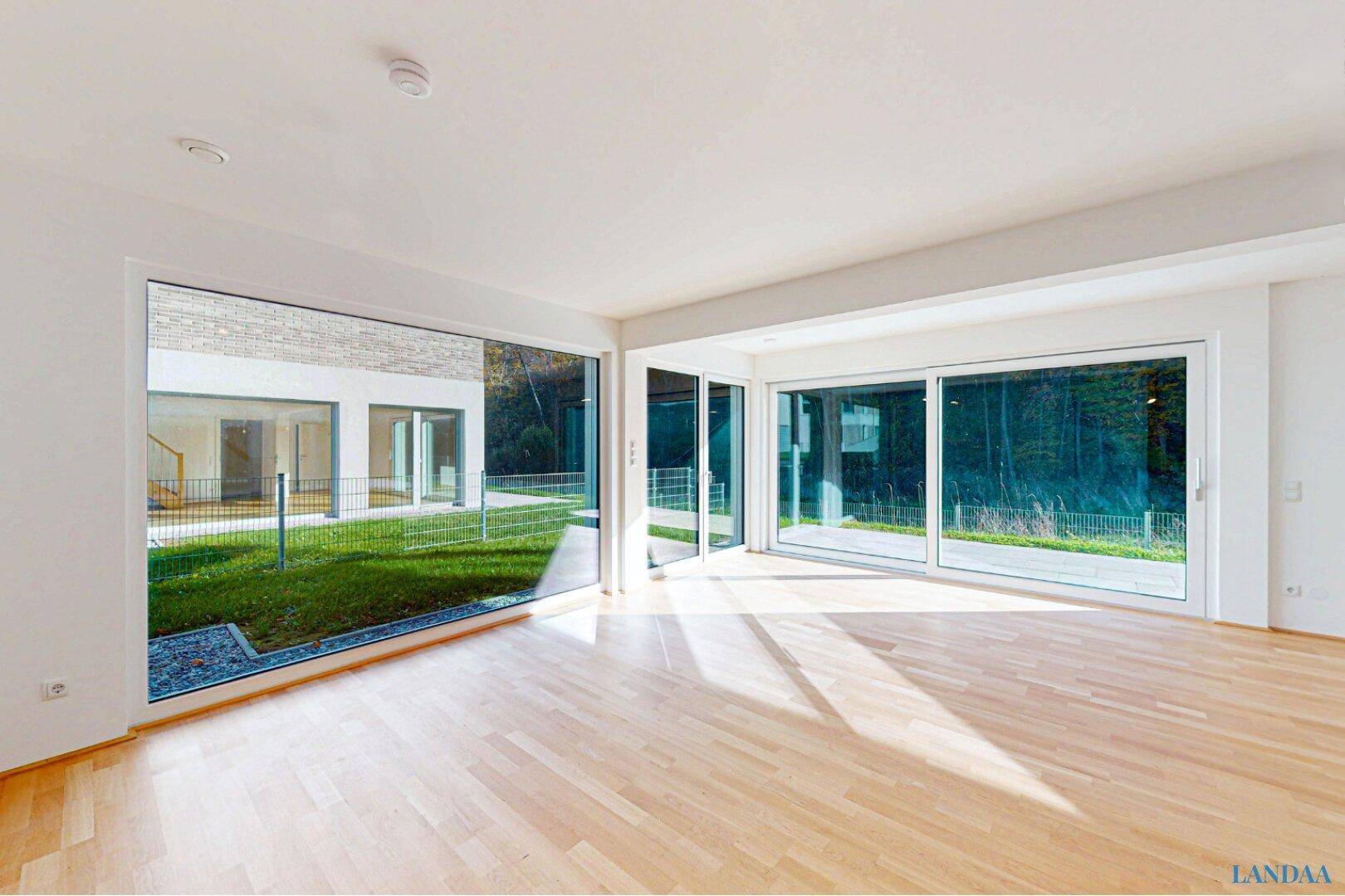 Heller, leerer Wohnraum mit großen Fenstern, Holzboden und Blick in den Garten.