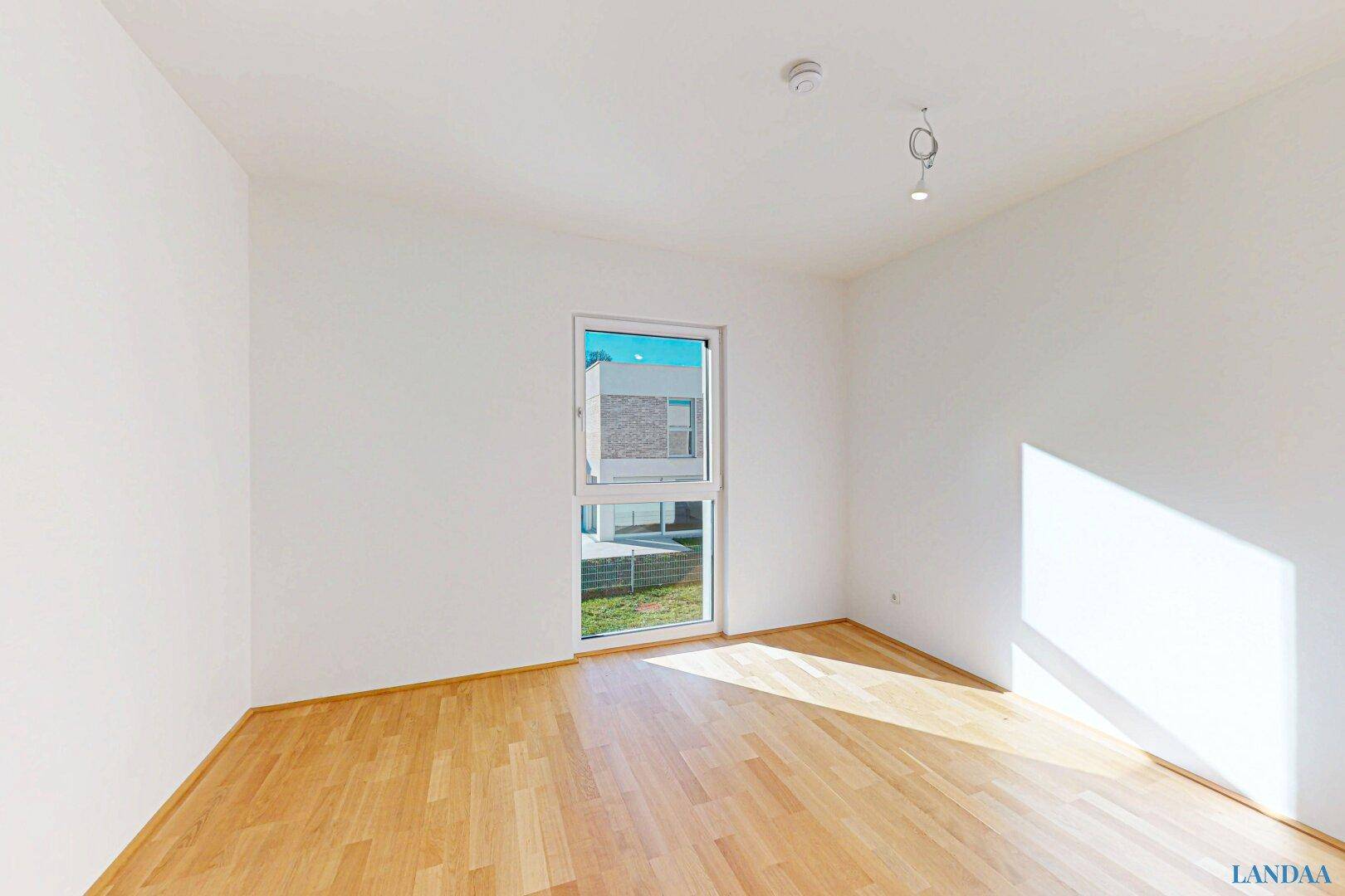 Leeres Zimmer mit hellem Holzboden und einem Fenster, das viel Tageslicht hereinlässt.