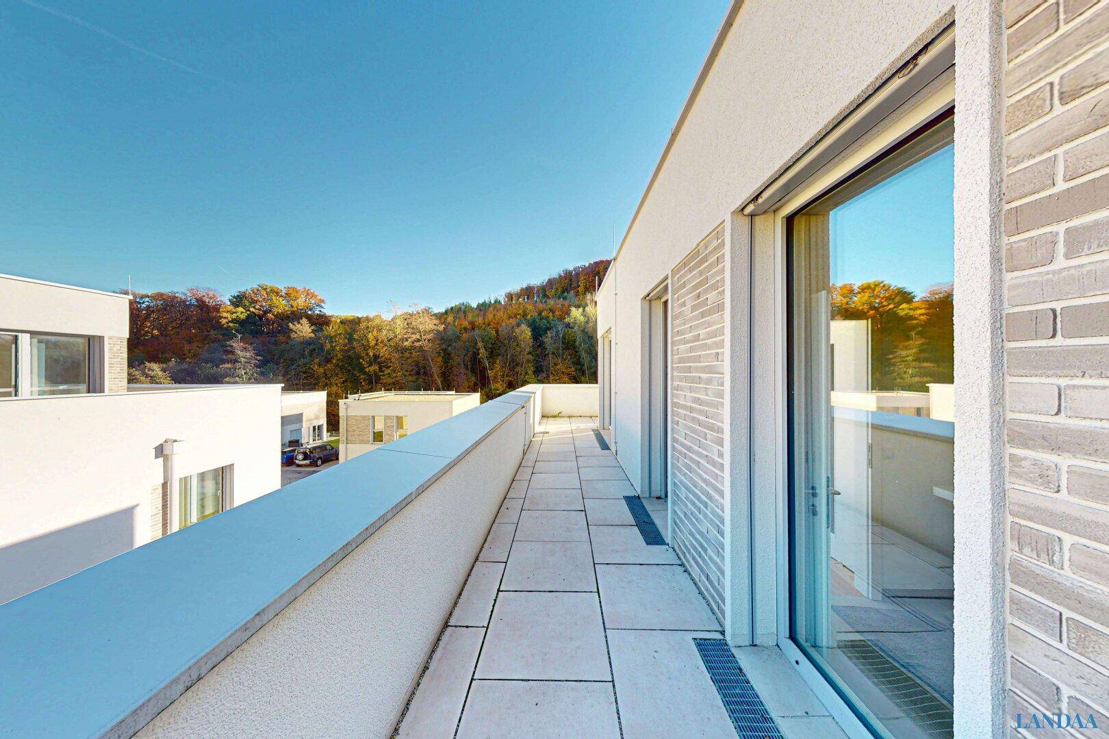 Langer, sonniger Balkon mit Fliesenboden und weitem Blick auf bewaldete Hügel.