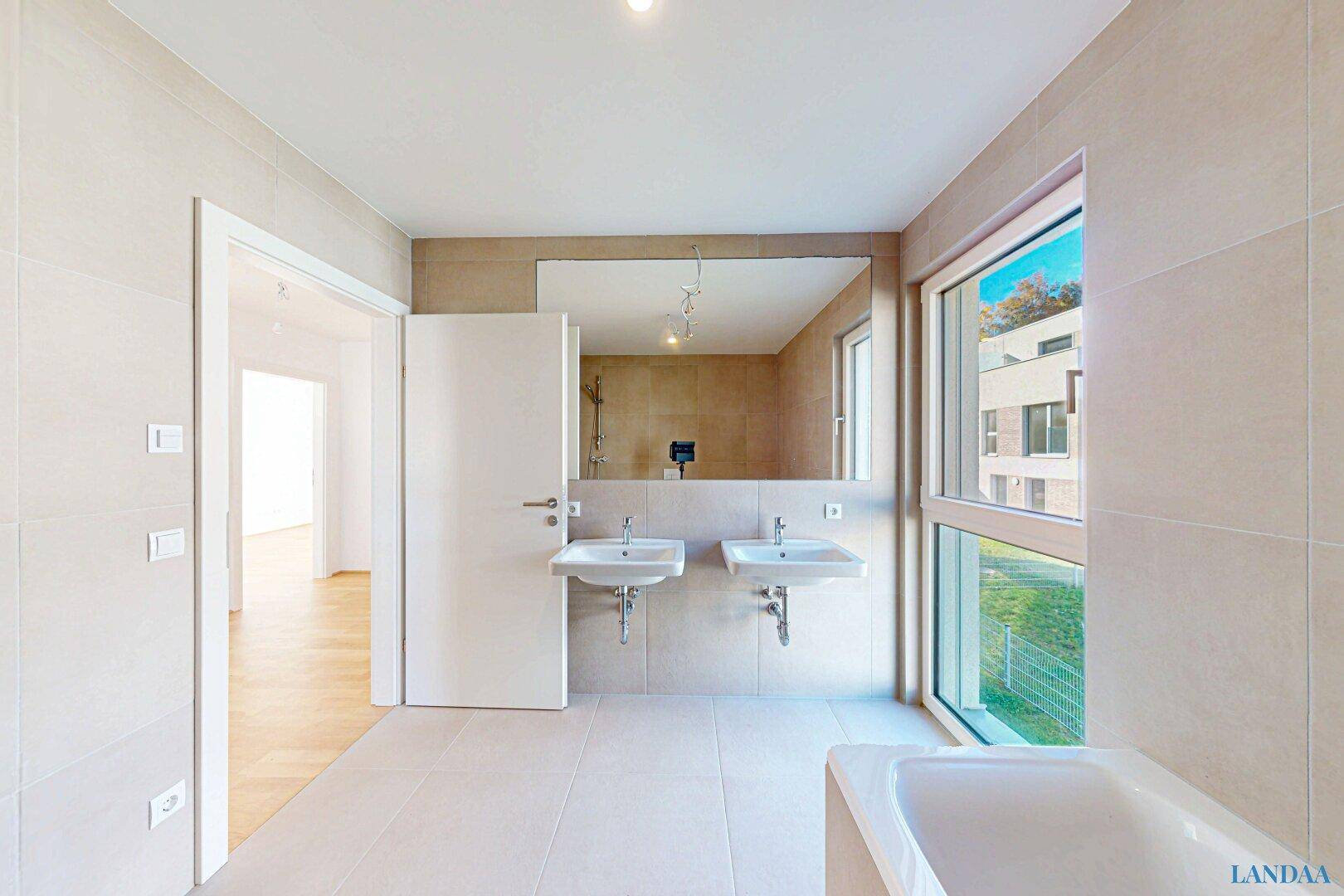 Modernes Badezimmer mit beigefarbenen Fliesen, Doppelwaschbecken und großer Badewanne am Fenster.