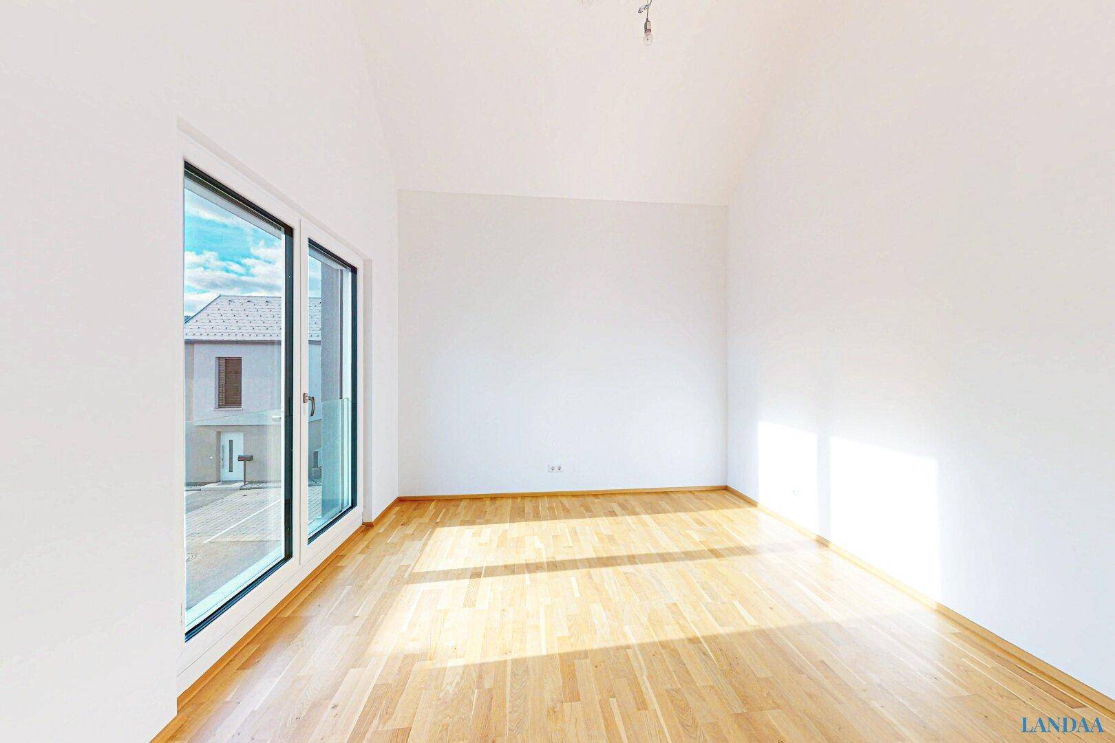 Heller, leerer Raum mit Holzboden und großer Fensterfront, die viel Sonnenlicht hereinlässt.