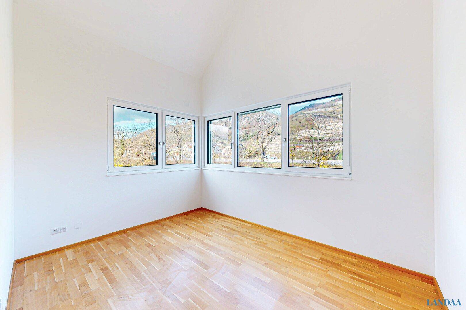 Geräumiger, heller Raum mit Holzboden, Dachschräge und Panoramafenstern mit Ausblick.