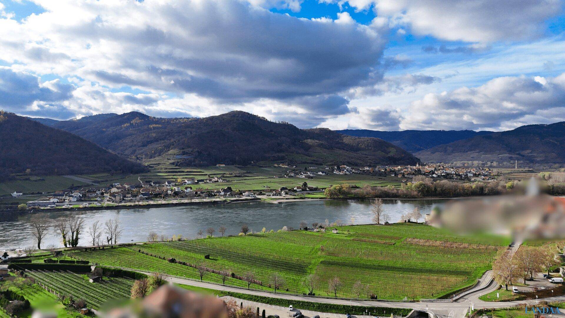 Weitläufige Landschaft mit Fluss, grünen Weinbergen und Bergen unter einem bewölkten Himmel.