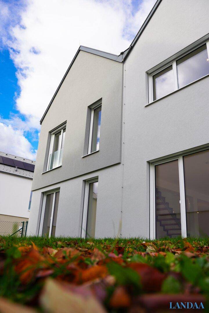 Moderne Hausfassade in hellem Grau, umgeben von Herbstlaub und blauem Himmel.