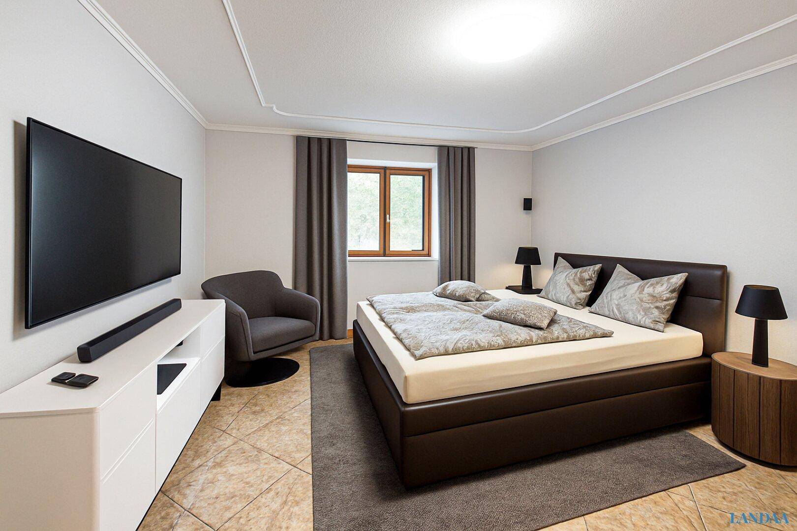 Gemütliches Schlafzimmer mit großem Doppelbett, Flachbildfernseher und beigen Fliesenboden.