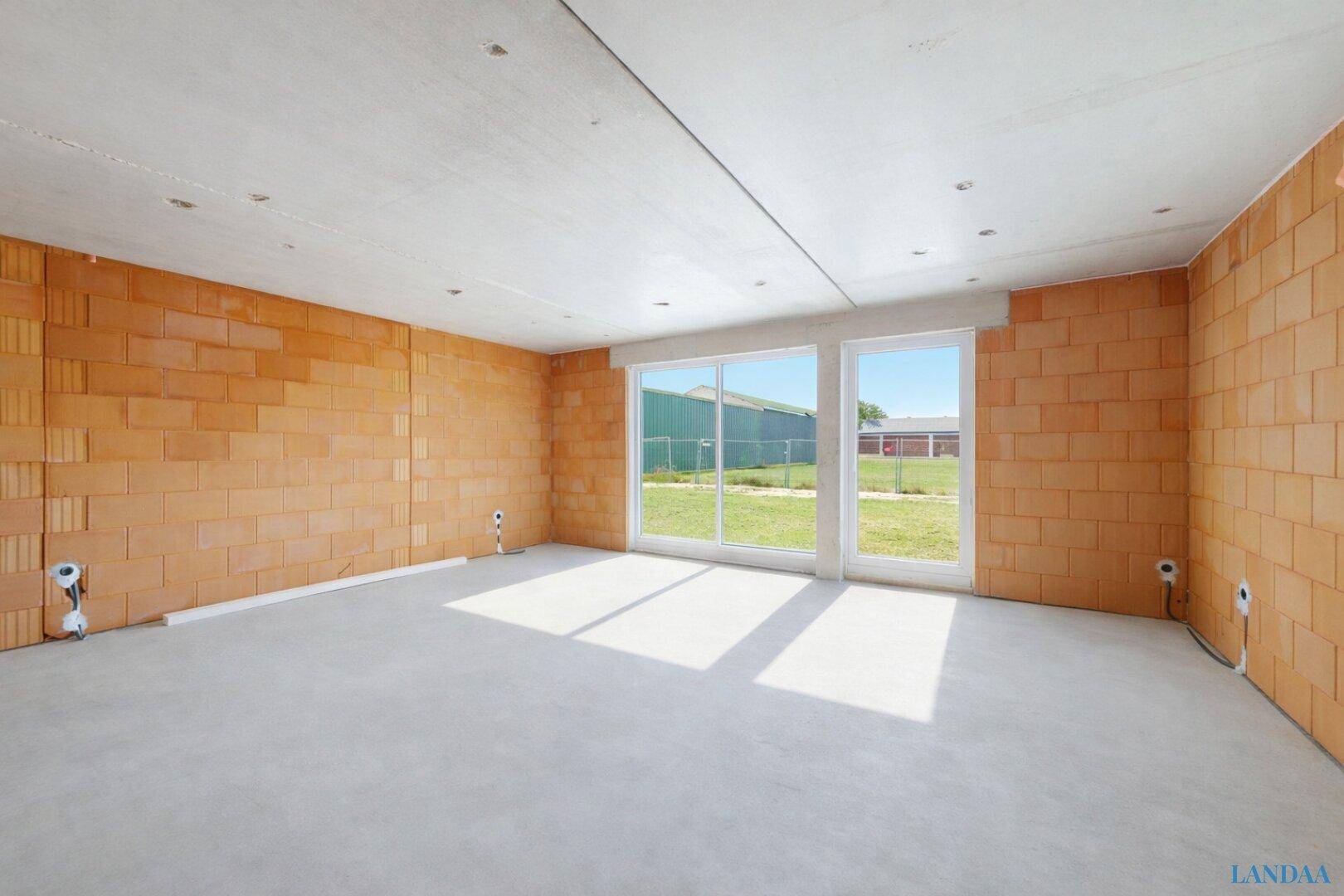 Innenraum im Rohbau mit Ziegelwänden, Betonboden und großen Fenstern mit Blick ins Grüne.