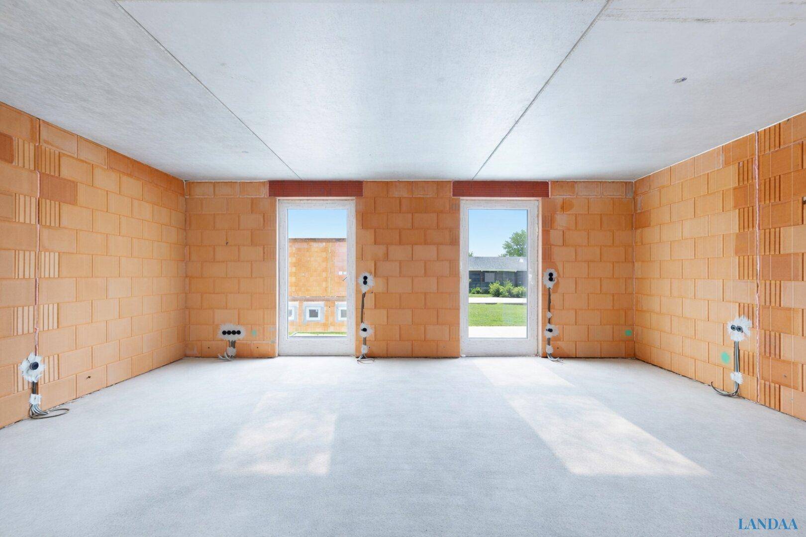 Innenraum im Rohbau mit Ziegelwänden, Betonboden und zwei Fenster- oder Türöffnungen.