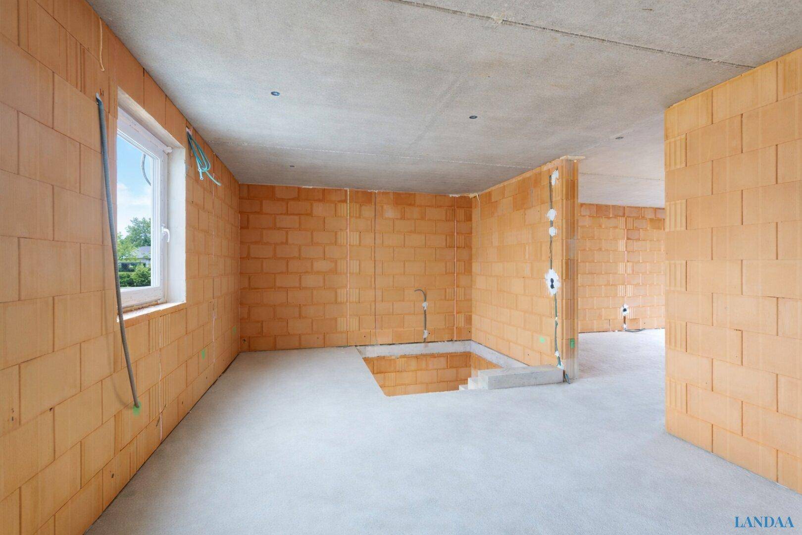 Innenraum im Rohbau mit Ziegelwänden, Betonboden, Fenster und einer Öffnung für eine Treppe.