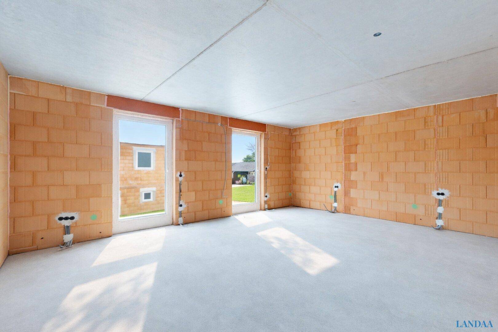 Innenraum im Rohbau mit Ziegelwänden, Betonboden und zwei Fenster- oder Türöffnungen mit Lichteinfall.
