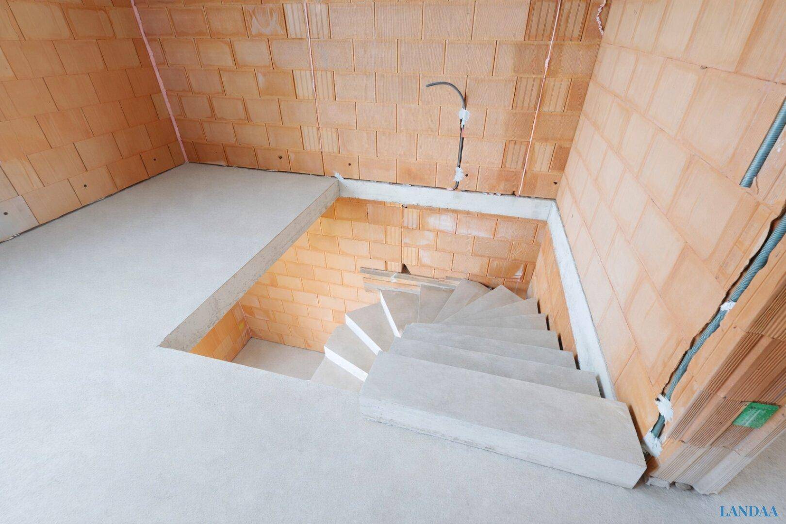 Innenraum im Rohbau mit Ziegelwänden, Betonboden und einer freitragenden Betontreppe.