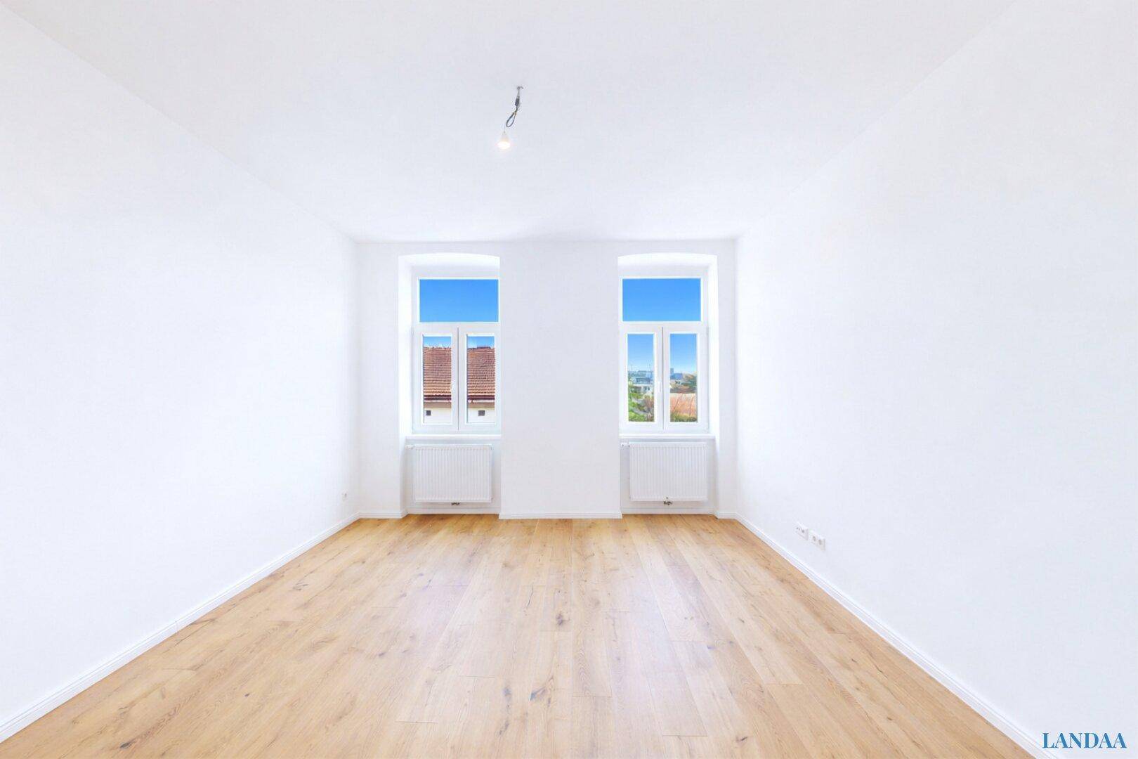 Geräumiges, leeres Zimmer mit hellem Holzfußboden und zwei großen Fenstern.