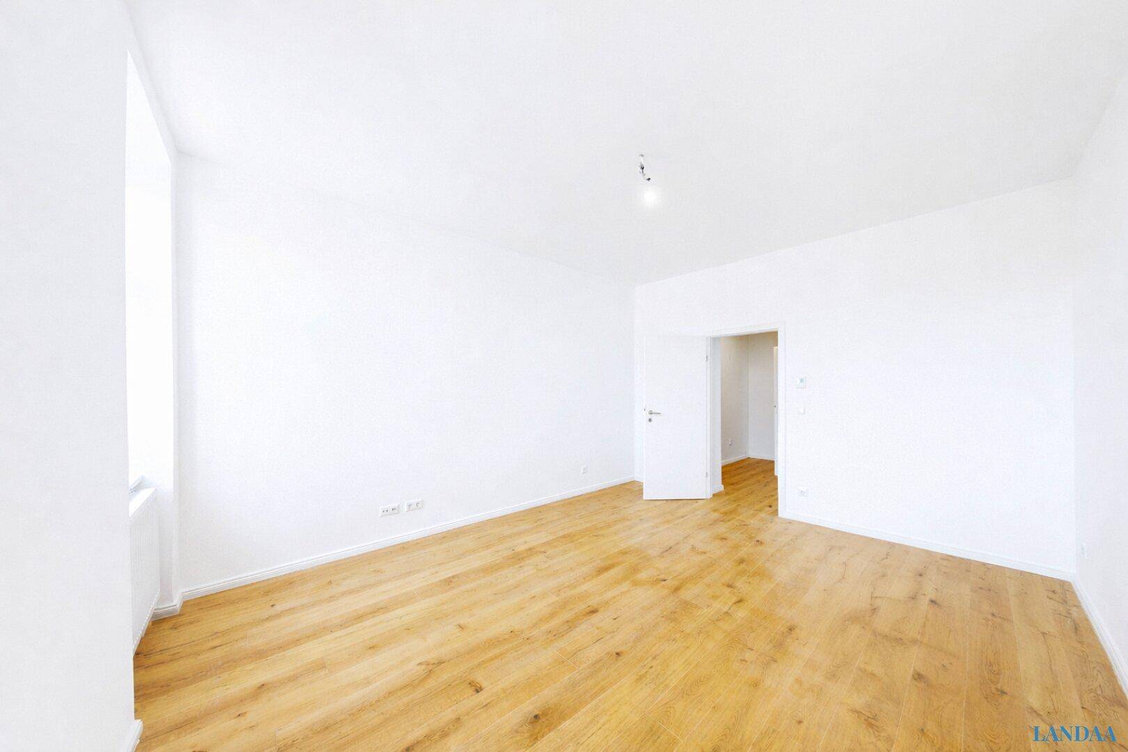 Helles, leeres Zimmer mit Holzfußboden und Zugang zu einem weiteren Raum.