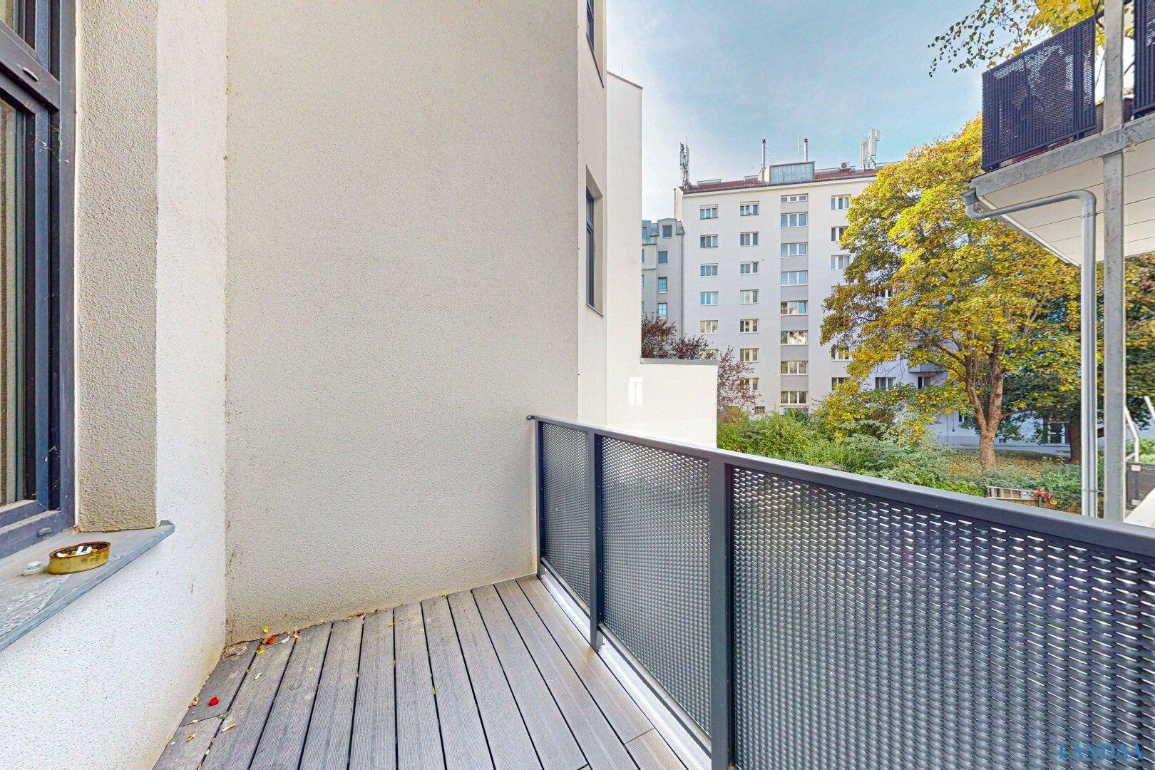 Kleiner Balkon mit Holzboden und Blick auf Bäume und umliegende Gebäude.