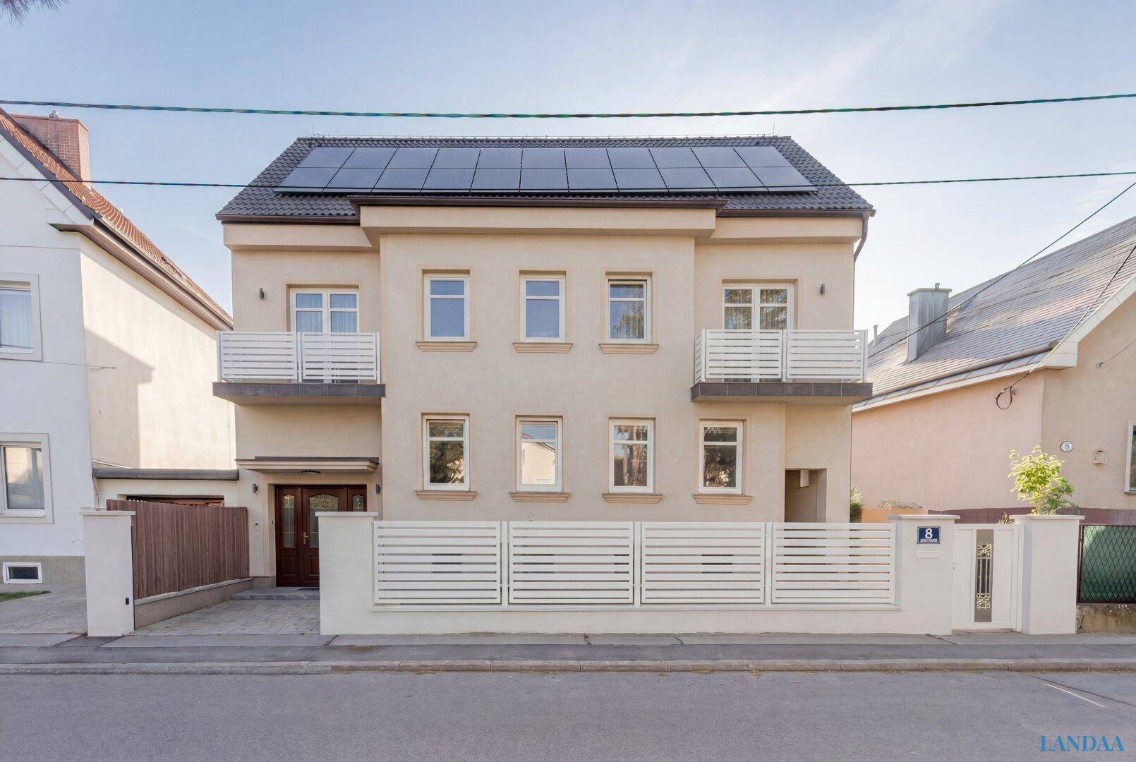 Fassade eines Mehrfamilienhauses mit Balkonen, Photovoltaikanlage auf dem Dach und modernem Zaun.