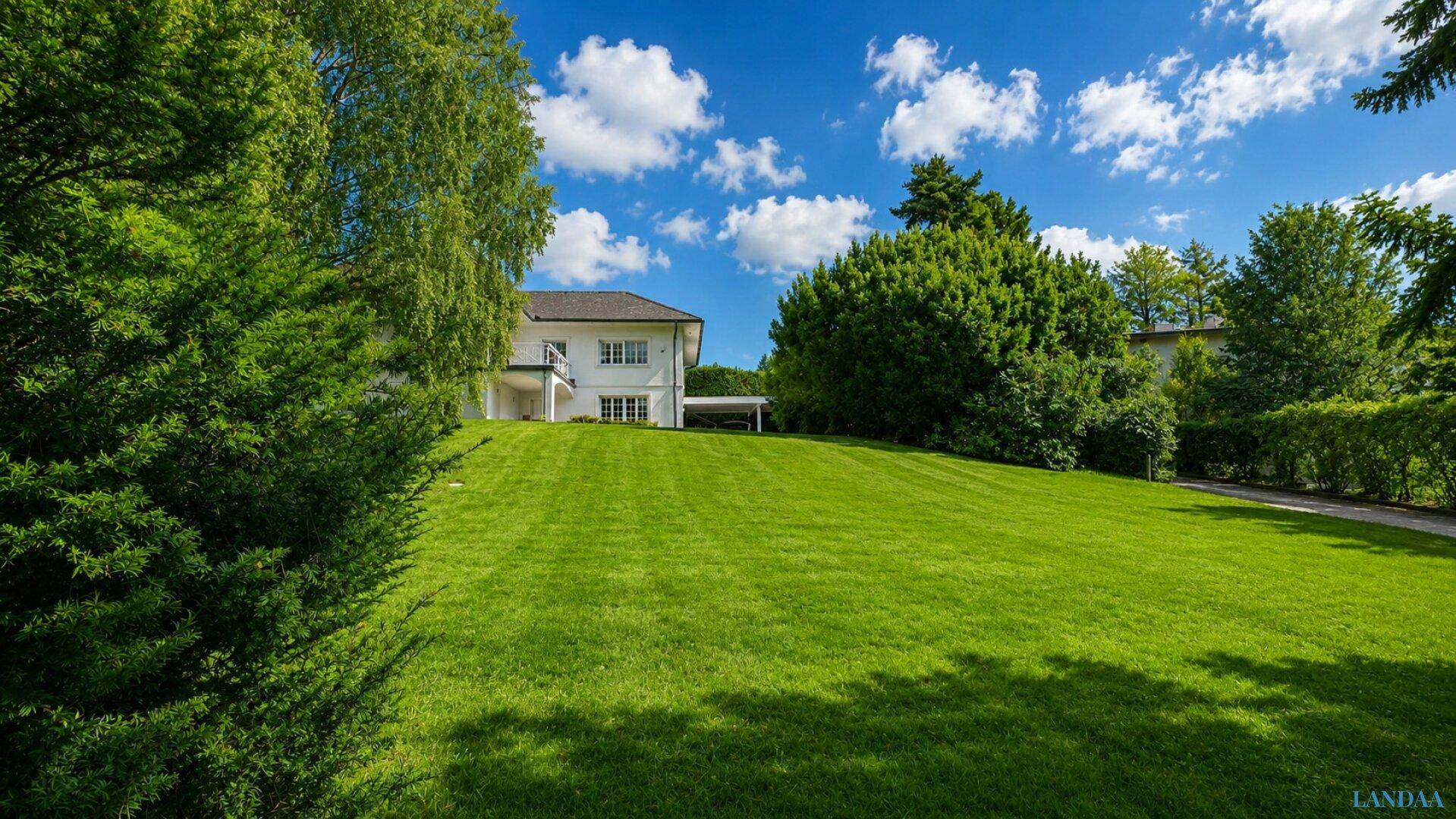 Weitläufiger, gepflegter Rasen mit Bäumen und dem Haus im Hintergrund unter blauem Himmel.