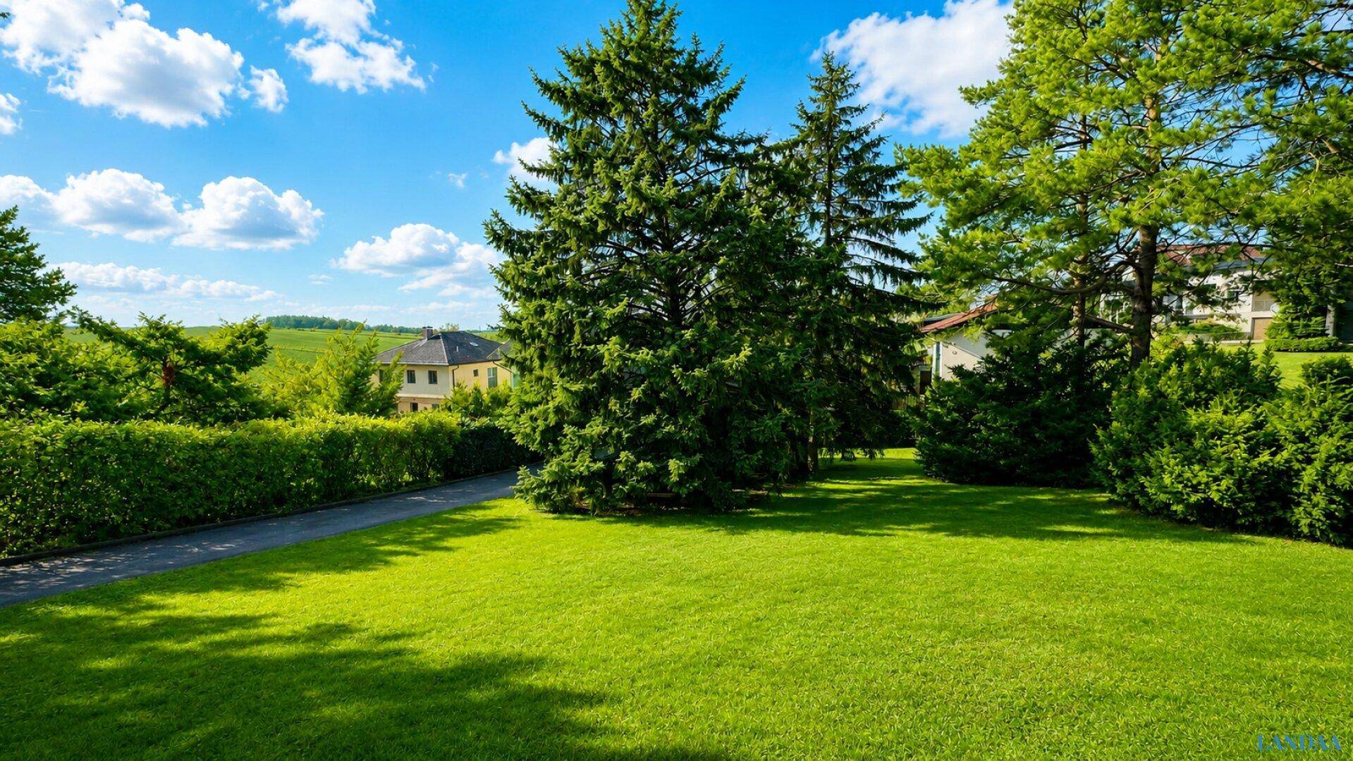 Weitläufige Rasenfläche mit Bäumen, Hecke und Blick auf die umliegende Landschaft.
