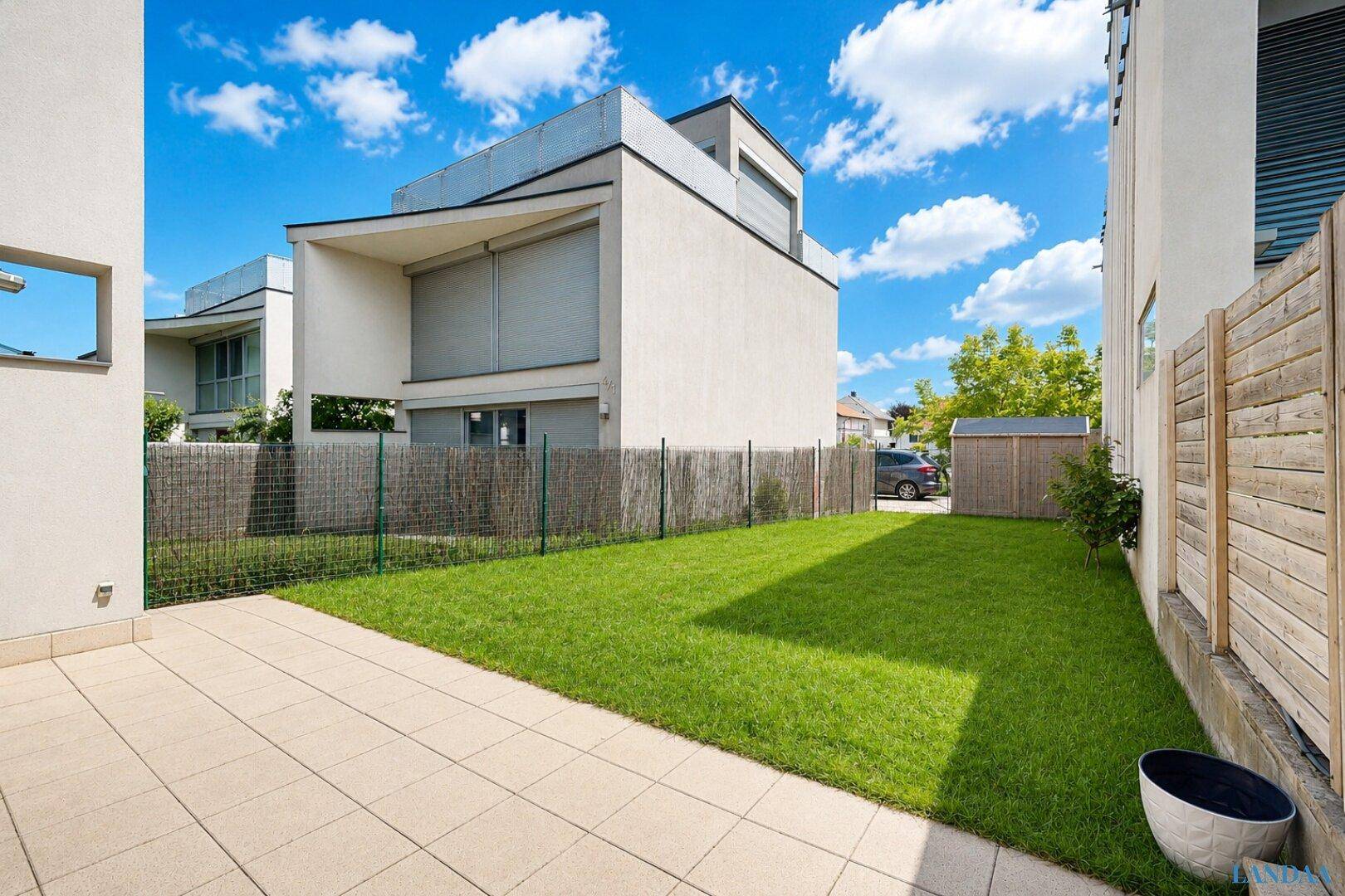 Gepflegter Garten mit Rasenfläche, gepflasterter Terrasse und Gartenhaus.