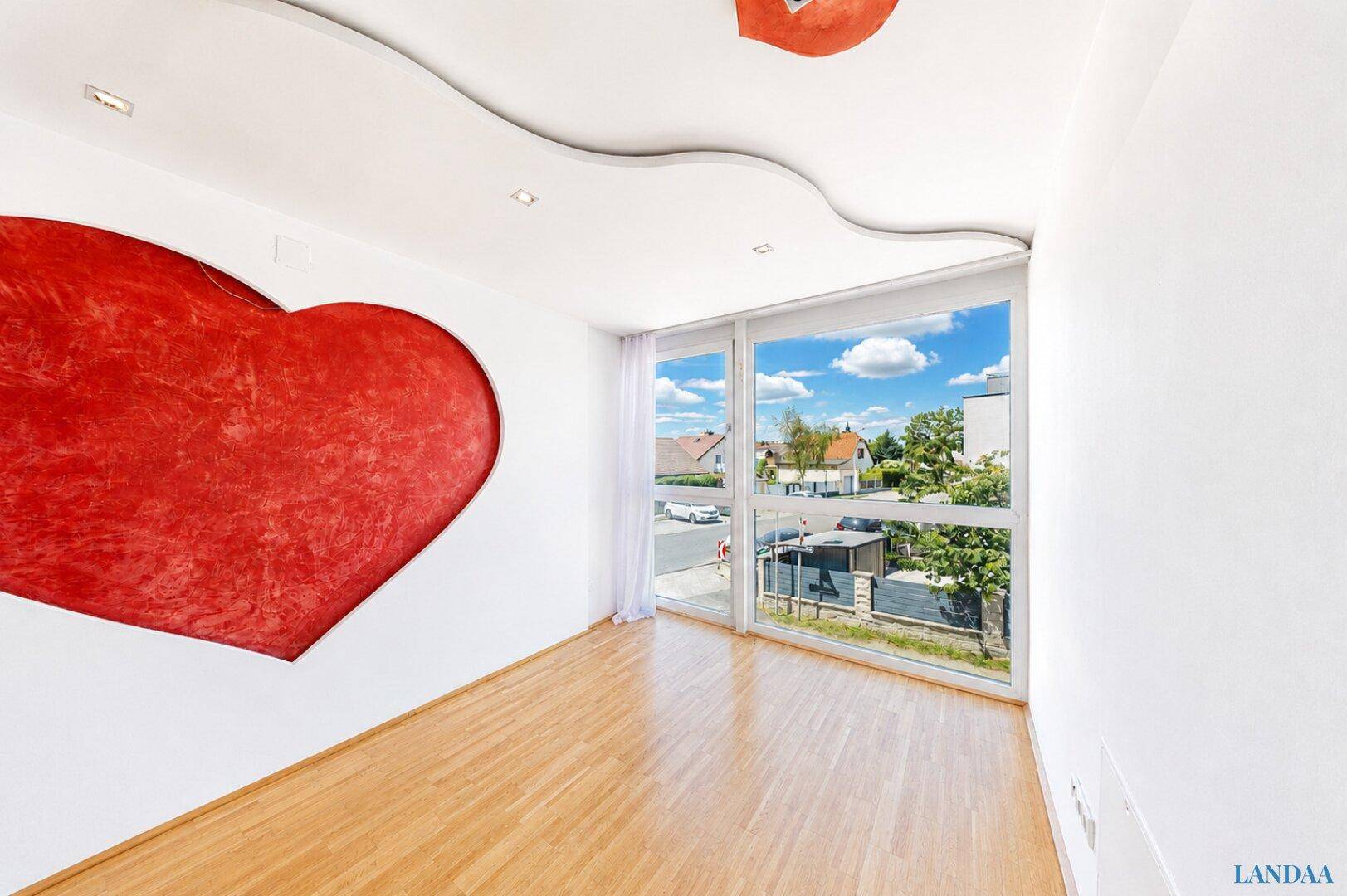 Helles Zimmer mit Holzboden, großem Fenster und auffälliger roter Herzdekoration.