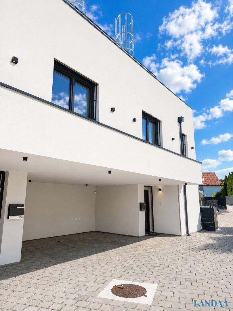 Moderne Gebäudefassade mit Carport und gepflasterter Fläche unter strahlend blauem Himmel.