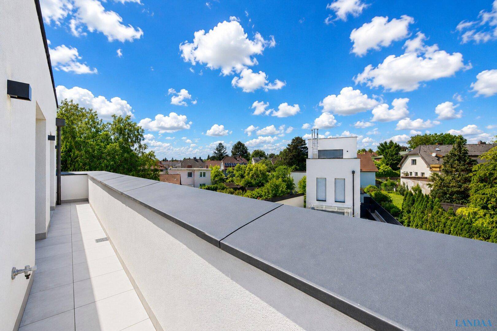 Großer Balkon mit weitem Blick über die Dächer und grüne Umgebung unter blauem Himmel.