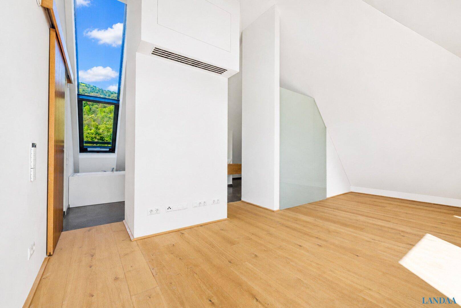 Heller Raum mit Holzboden, schrägen Wänden und einem Fenster, das einen Blick ins Grüne bietet.