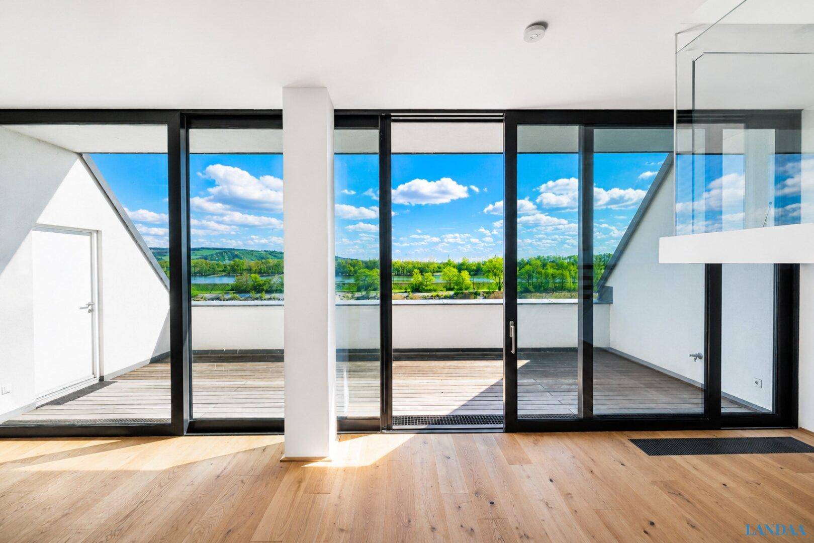 Heller Wohnbereich mit Holzboden und großen Glasfronten, die Zugang zu einer Terrasse mit Seeblick bieten.