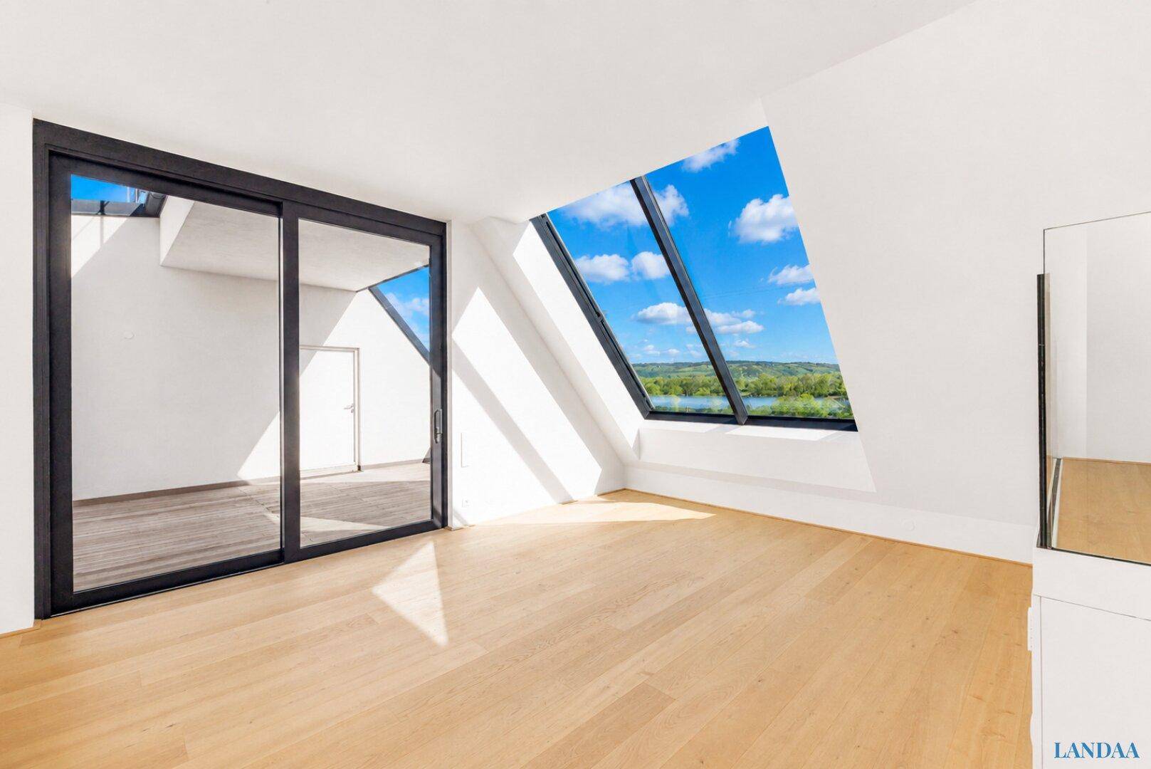 Heller, leerer Raum mit Holzboden, schrägen Wänden und großen Fenstern mit Blick auf die Natur.