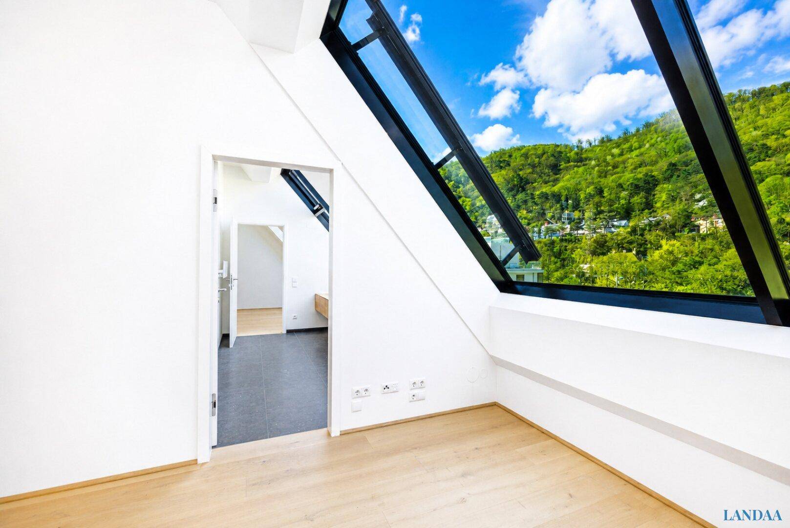 Heller Raum mit Holzboden, schrägen Wänden und einem großen Fenster mit Blick auf bewaldete Hügel.