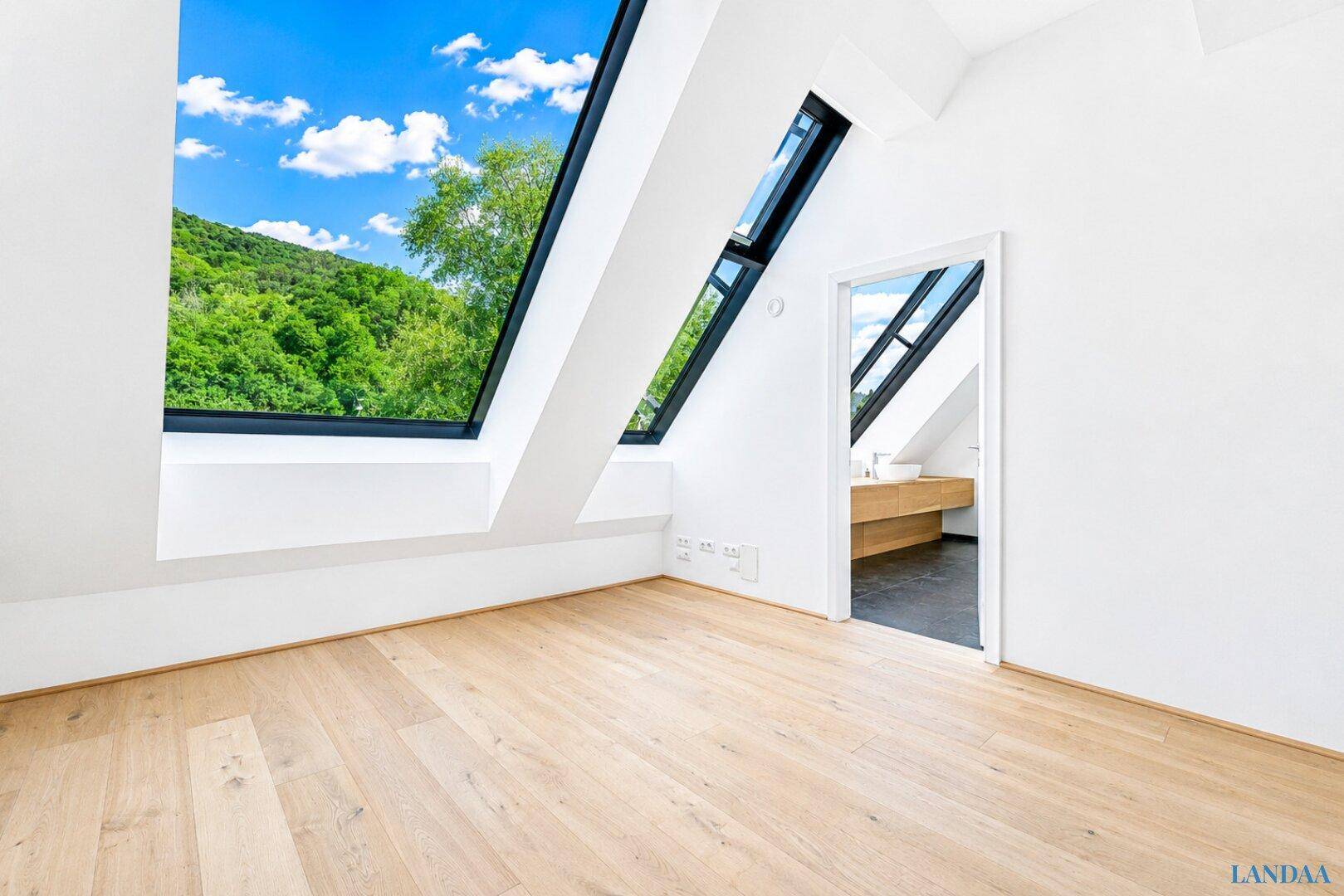 Heller Raum mit Holzboden, schrägen Wänden und einem Fenster, das einen Blick ins Grüne bietet.
