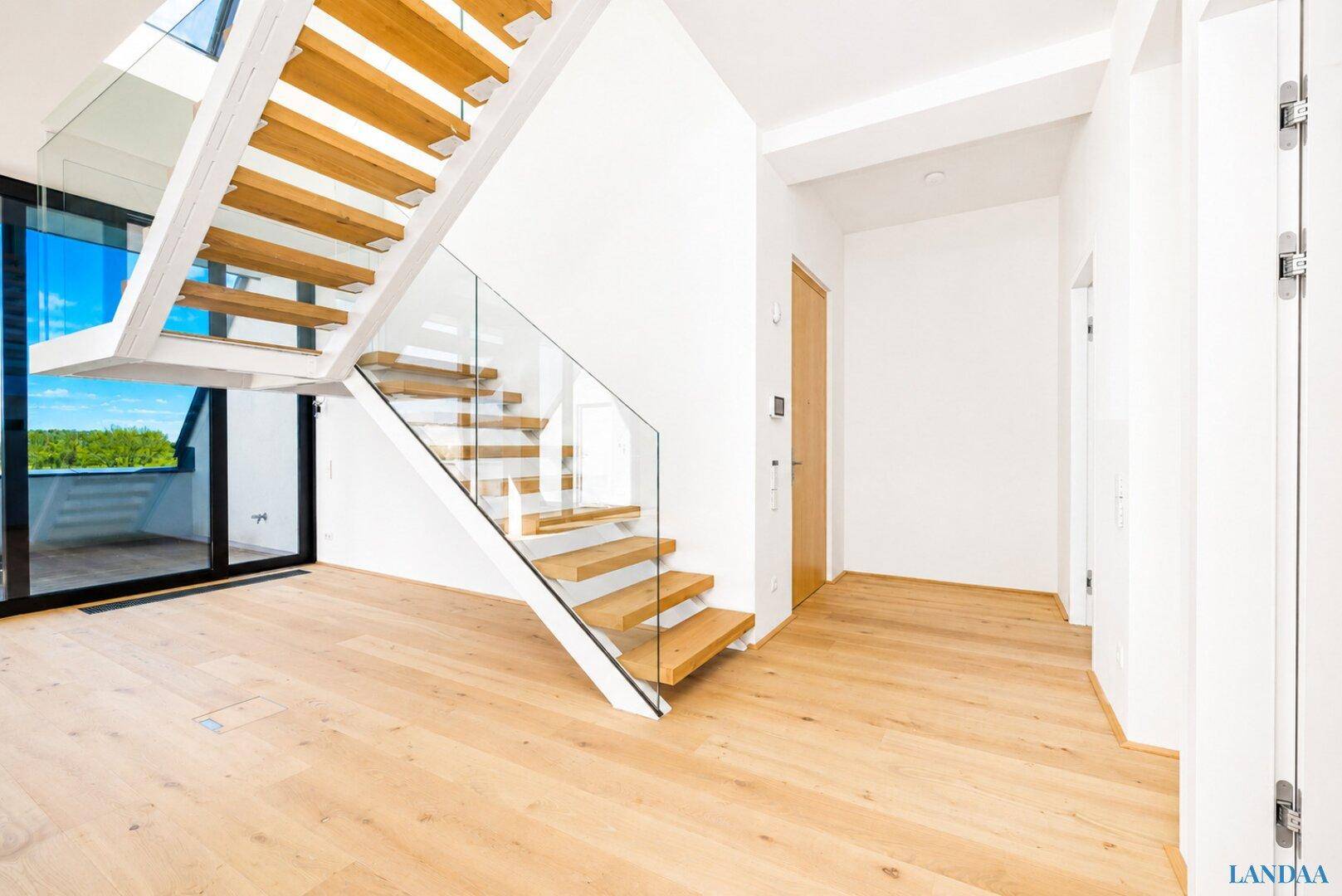 Heller Eingangsbereich mit moderner Holztreppe und Glasgeländer, die zu den oberen Etagen führt.