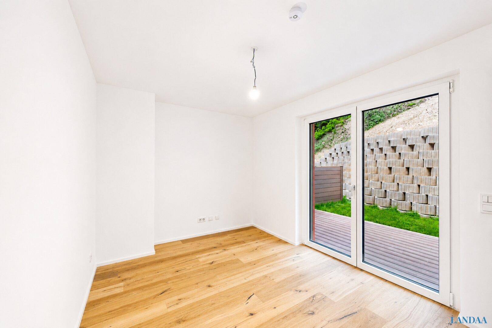 Heller, leerer Raum mit Holzboden und großen Fenstern, die auf eine begrünte Terrasse führen.