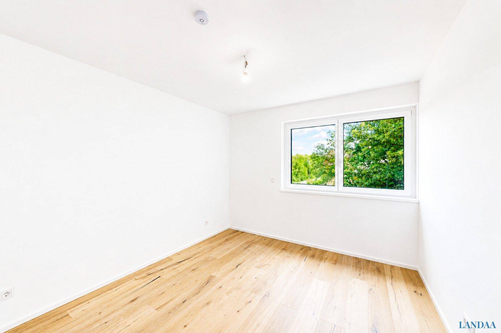 Heller, leerer Raum mit weißen Wänden, Holzboden und Fenster mit Blick ins Grüne.
