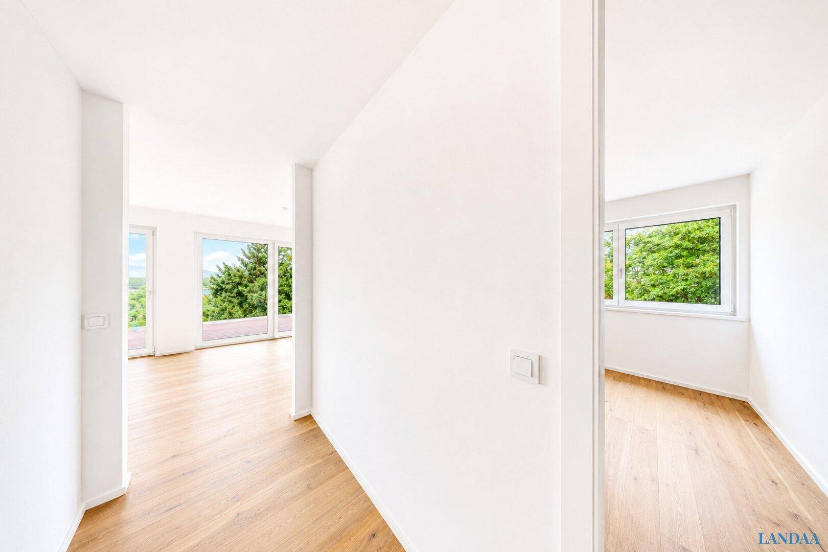 Heller Flur mit Holzboden und weißen Wänden, der zu Räumen mit großen Fenstern führt.