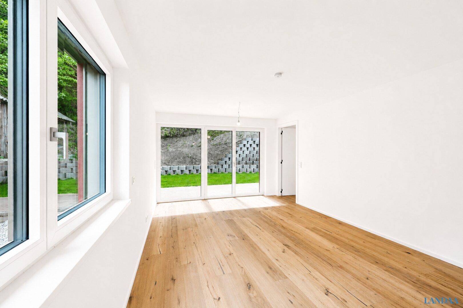 Heller, leerer Raum mit Holzboden und großen Glasfronten zum Garten.