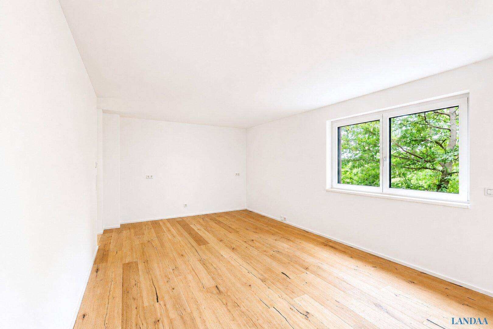 Leeres Schlafzimmer mit Holzboden und Fenster mit Blick ins Grüne.