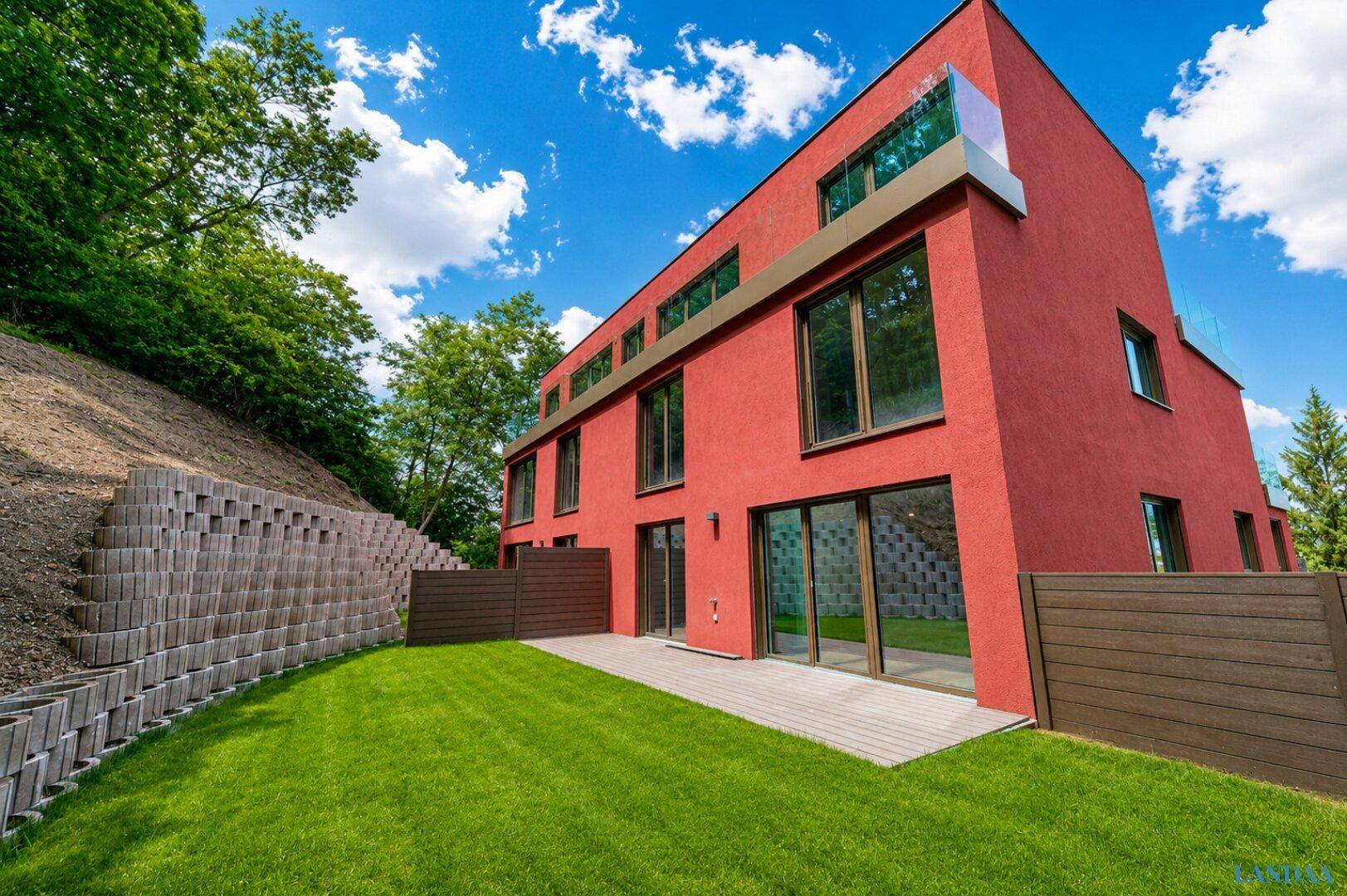 Rückseite des modernen Gebäudes mit Terrasse, Rasenfläche und Hangbegrünung.